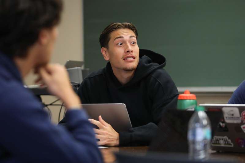 a man in a black hoodie sitting at a table with a laptop