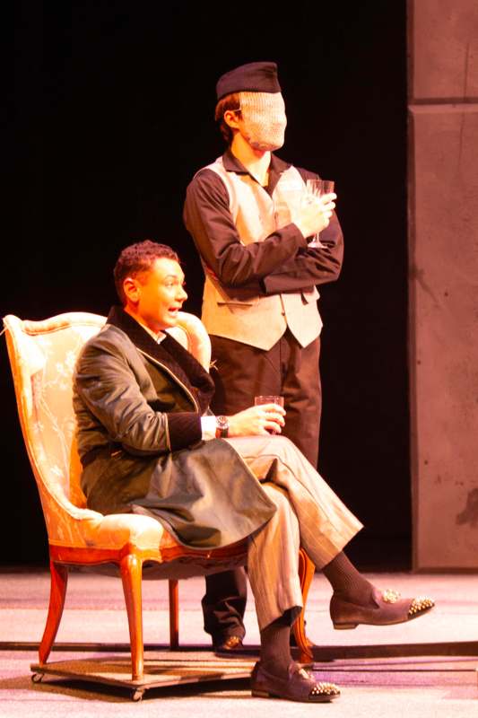 a man in a mask holding a glass of wine next to a man in a chair