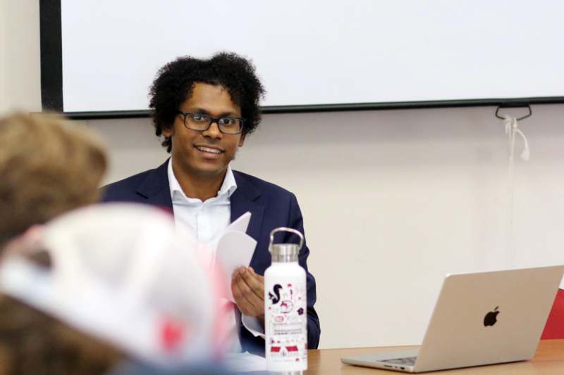 a man in a suit and glasses sitting at a table with a laptop and a white board