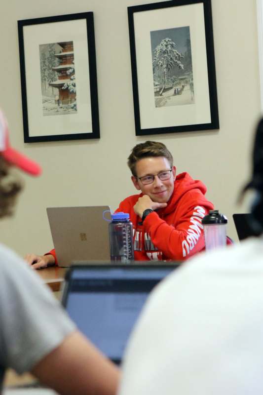 a man in a red hoodie sitting at a table with a laptop