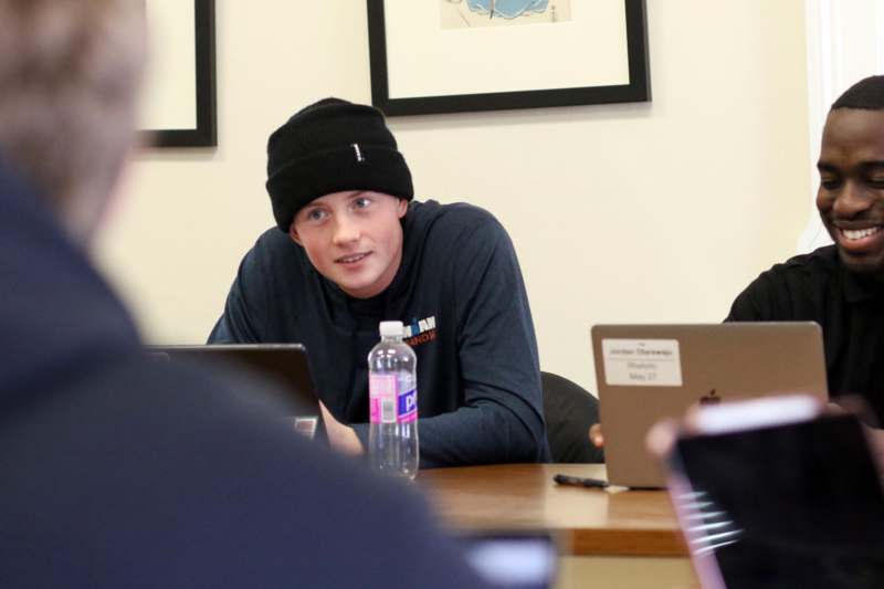 a man in a beanie sitting at a table with laptops and water bottle