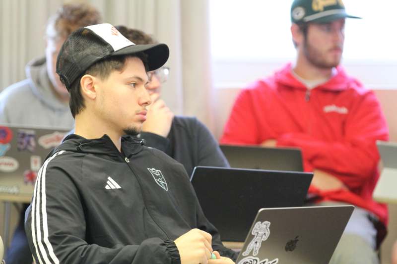 a man in a black jacket and baseball cap sitting in front of a laptop