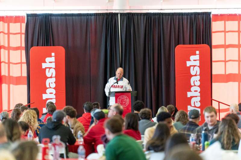 a man speaking at a podium with a crowd of people seated at tables