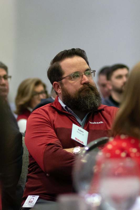 a man with a beard and glasses sitting in a room with other people
