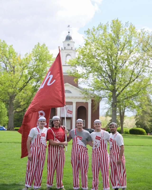 Five Wabash students wearing red and white stripped overalls stand in front of Pioneer Chapel holding a Wabash scarlet banner